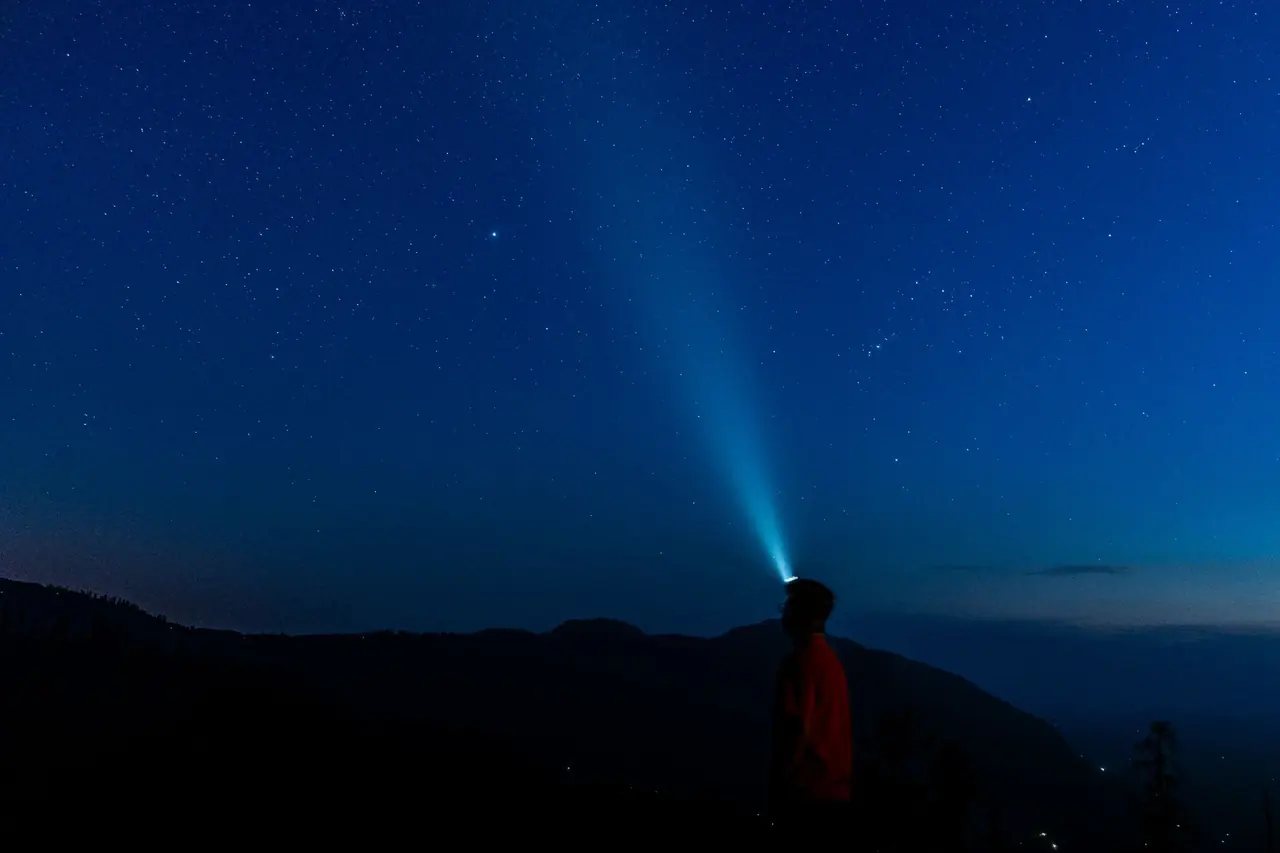 Silhouette d'une personne avec lampe frontale dont le faisceau éclaire le ciel étoilé